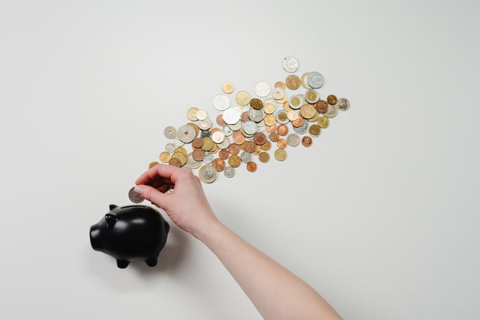 A black piggy bank with a cloud of coins above with against a white background, with a hand putting more coins into the piggybank
