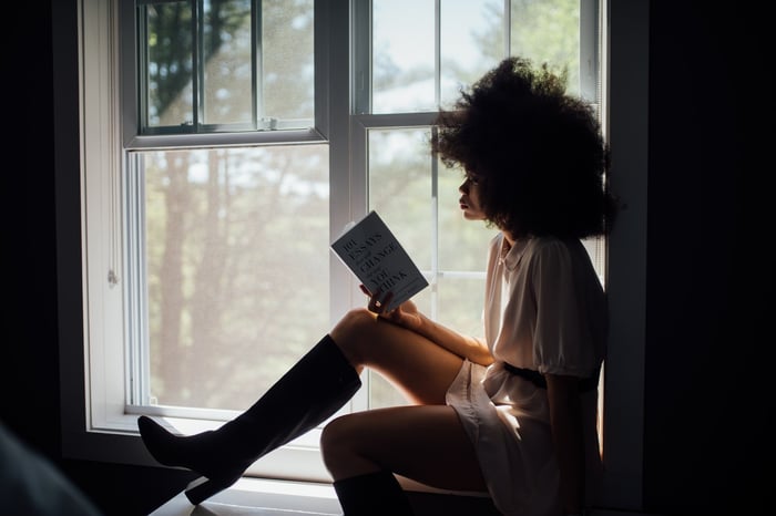 Una mujer leyendo un libro junto a un alféizar de la ventana. 