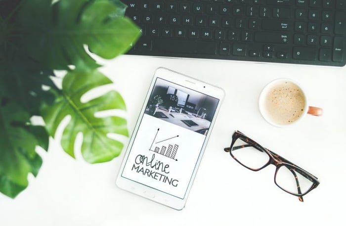 Mesa branca com uma planta, óculos, xícara de café e um tablet com a frase "online marketing" na tela