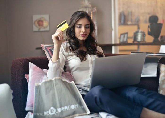 Mulher sentada no sofá olhando para o notebook, com um cartão de crédito na mão e sacolas de compras ao redor
