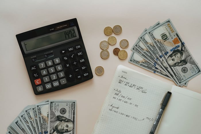 Dinheiro, calculadora e caderno com anotações sobre uma mesa branca
